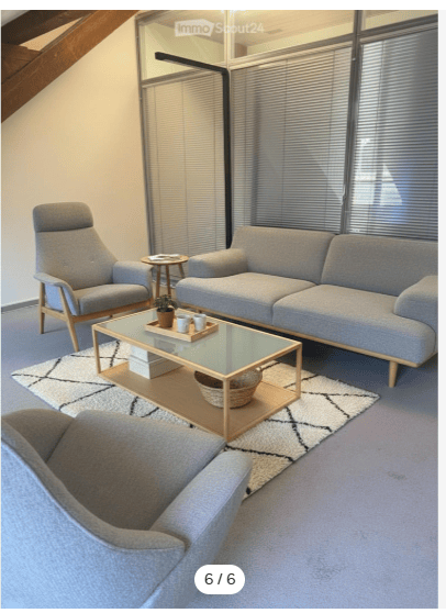 Modern living room with grey sofa, armchair, glass coffee table, side table, and small plant on the coffee table. The room features large windows with blinds and a patterned rug on the floor.