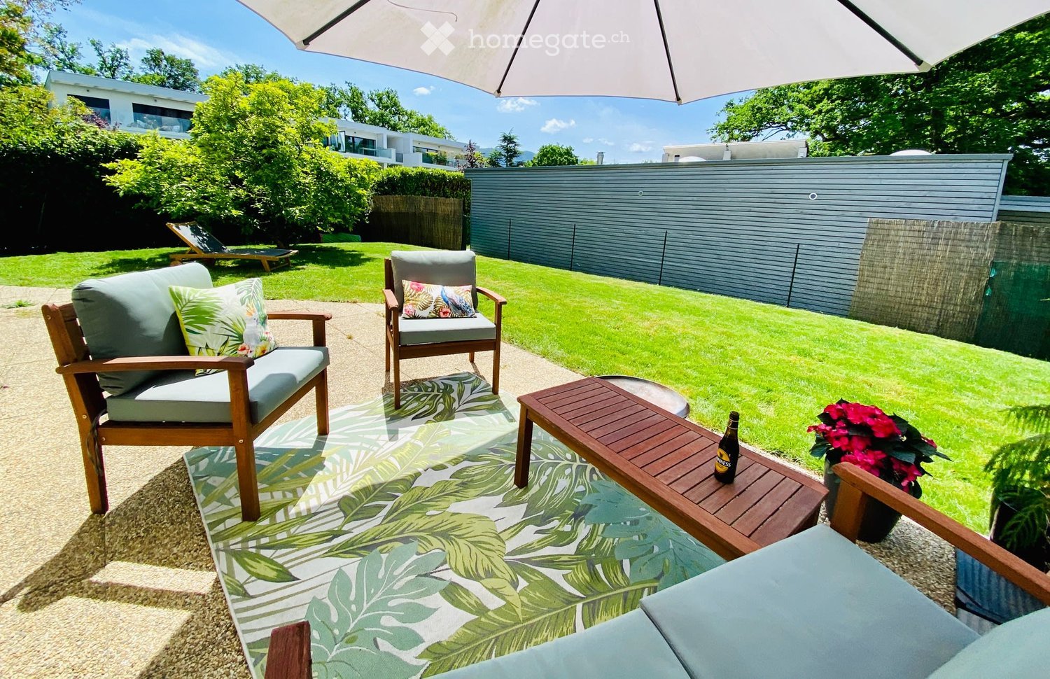 A well-maintained garden with a seating area featuring a sofa, chairs, a coffee table, and a large umbrella providing shade. The area is surrounded by a neatly trimmed lawn, trees, and a modern building in the background.