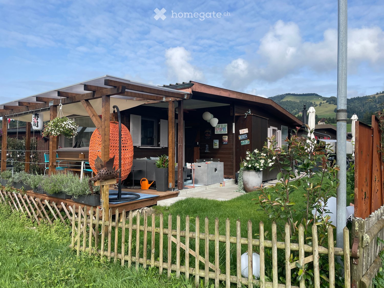A modern wooden house with a covered patio area, outdoor seating, and a well-maintained garden with a wooden fence.
