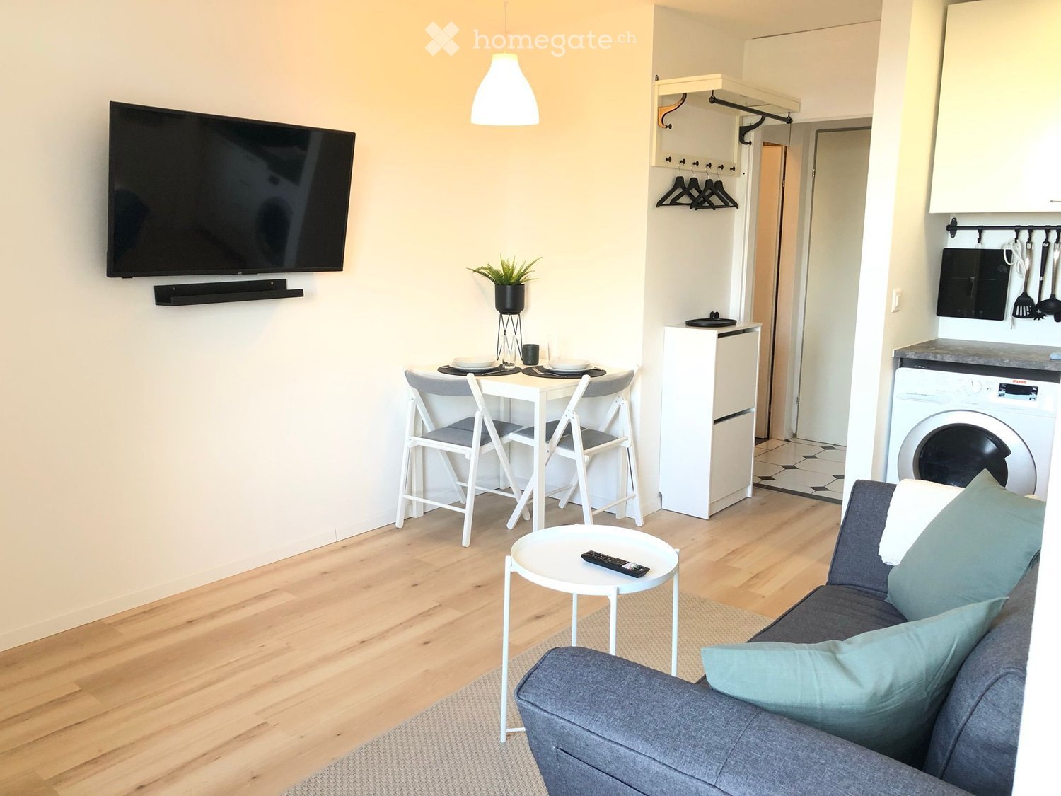 A modern, compact living space with a sofa, small round table, TV, dining area, and a small kitchen area with a washing machine.