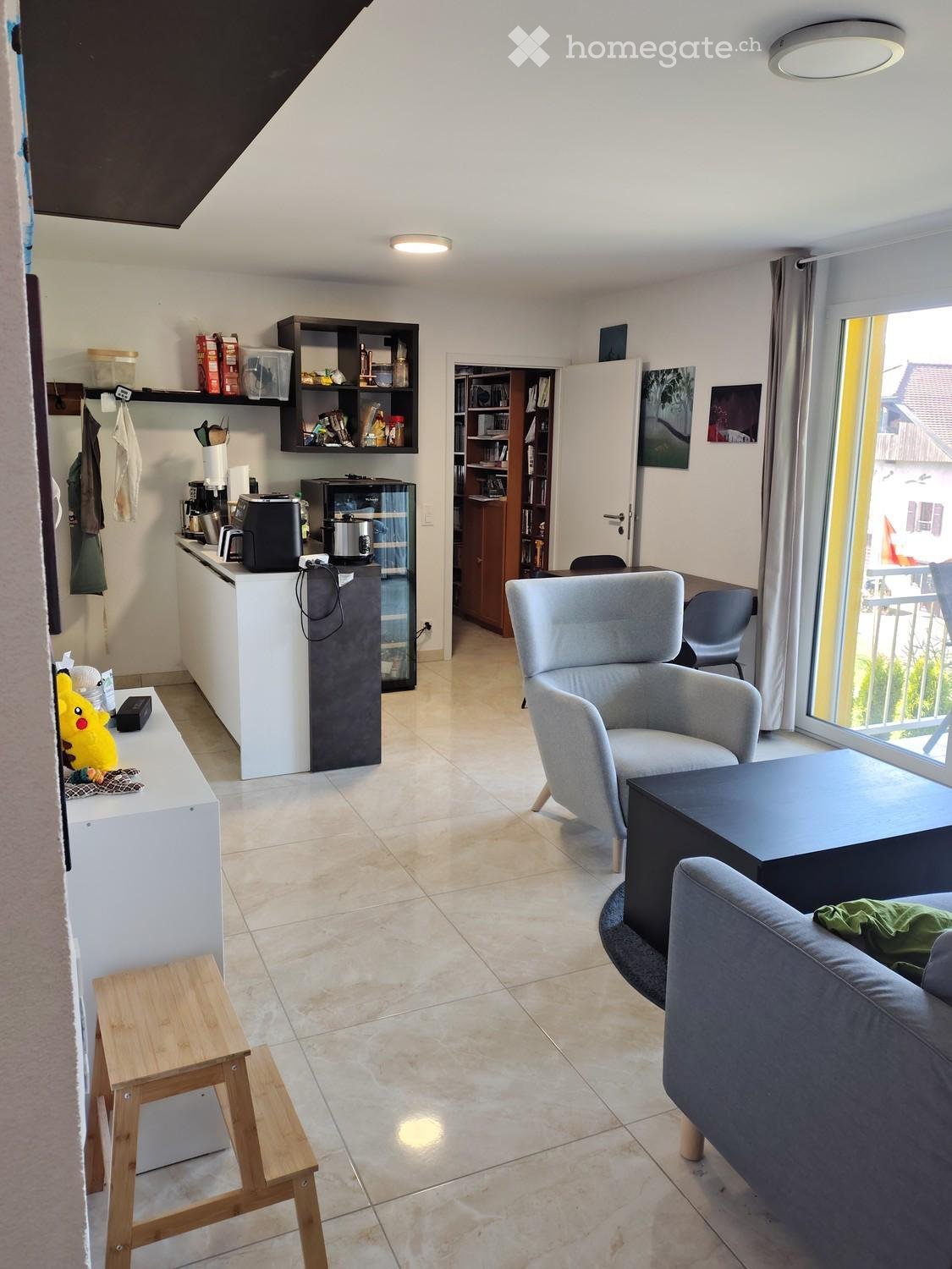 Modern living room with a grey sofa, armchair, coffee table, and a small kitchen area with a refrigerator and coffee maker. The space features tiled flooring, large windows, and a bookshelf with various items.