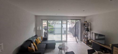Modern living room with large sliding glass doors leading to a balcony, grey sectional sofa with orange throw pillows, coffee table, floor lamp, TV stand, and a small shelf with decorative items.