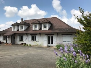 Two-story white house with a brown tiled roof, multiple dormer windows, and a garage. The house is surrounded by a driveway and some purple flowers in the foreground.