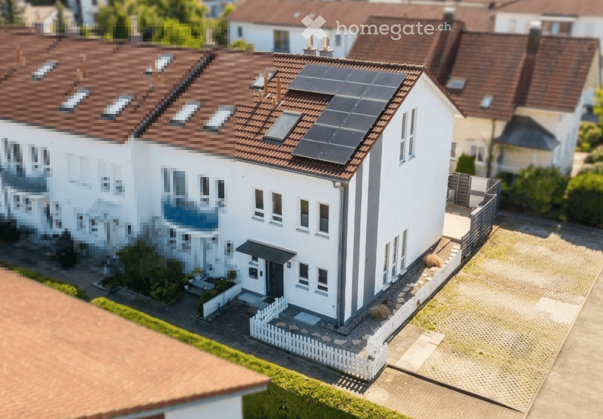 A modern two-story house with a white exterior, brown tiled roof, multiple windows, and solar panels on the roof. The house features a small front yard with a white picket fence and a paved driveway.