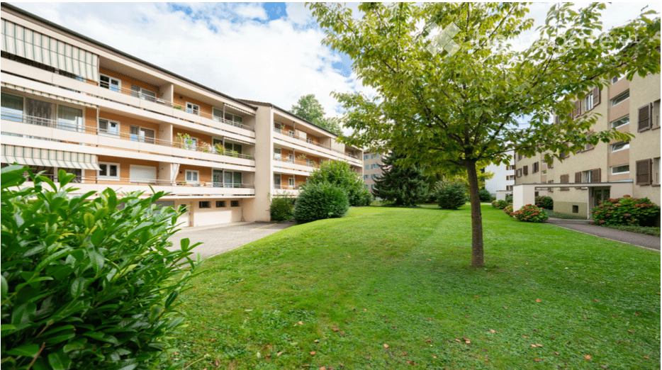 Well-maintained garden area with green grass, trees, and shrubs, surrounded by multi-story apartment buildings.