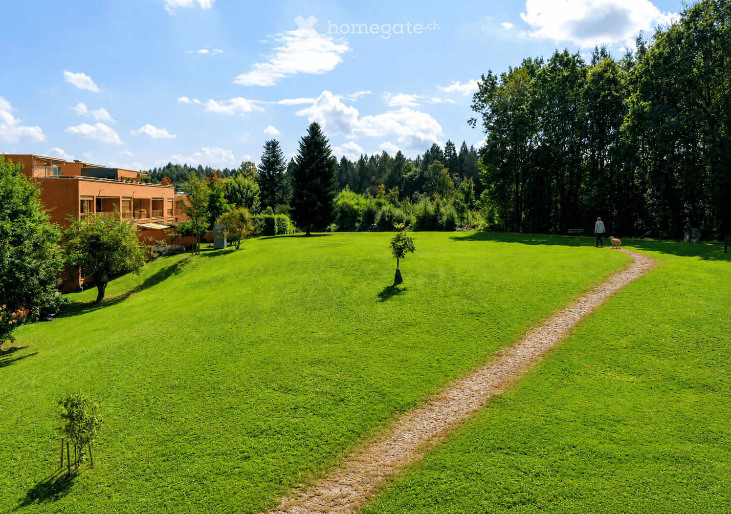 A large, well-maintained green lawn with a pathway leading to a building, surrounded by trees and shrubs under a partly cloudy sky.