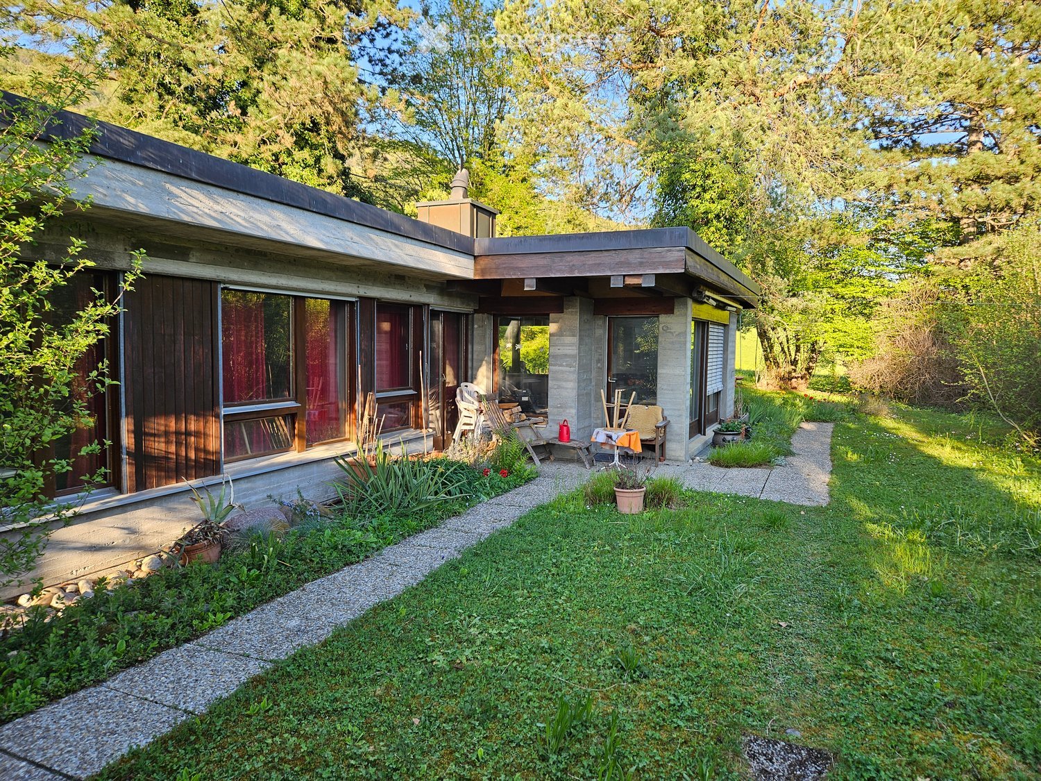 Modern single-story house with large windows, wooden paneling, and a small patio area with outdoor furniture.
