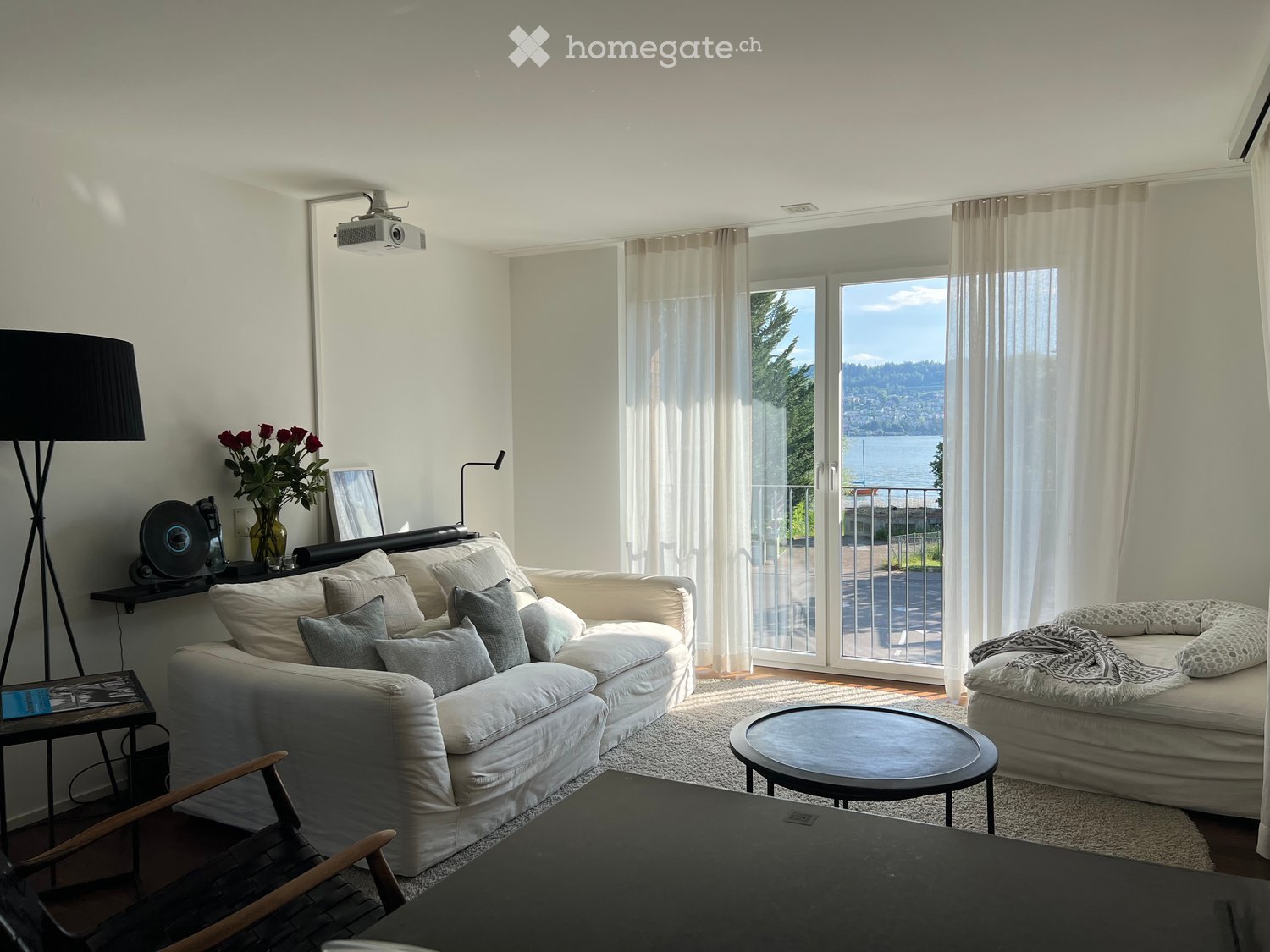 Bright living room with large windows and a view, featuring a white sofa, coffee table, ottoman, and a wooden chair.