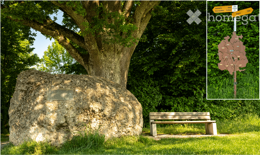 A large stone monument stands prominently in a grassy area, surrounded by lush greenery and trees. A wooden bench is positioned nearby, and a directional signpost is visible in the upper right corner, indicating nearby locations.