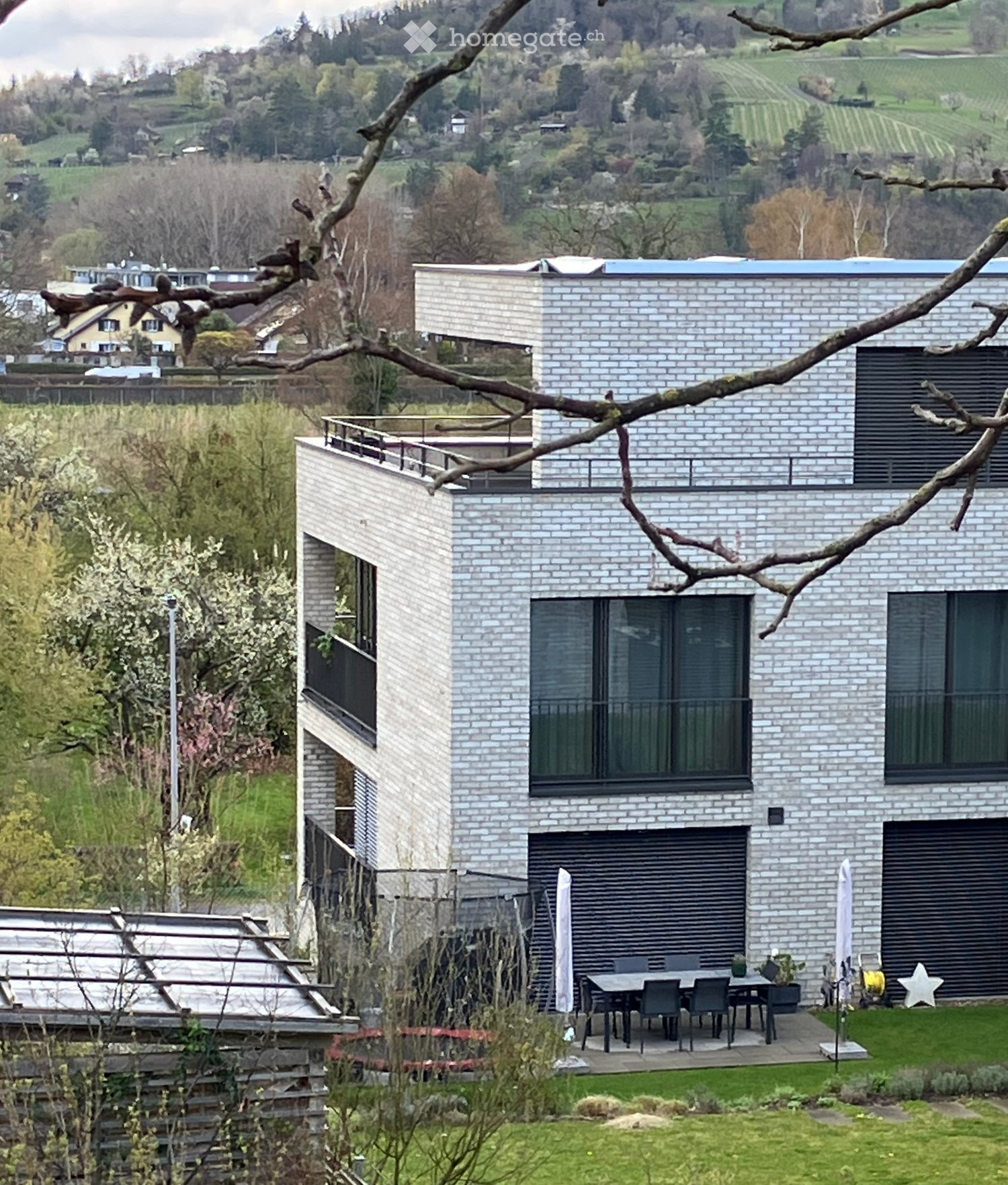 Modern two-story house with white brick facade, large windows, balconies, and outdoor dining area surrounded by greenery and scenic hillside view.