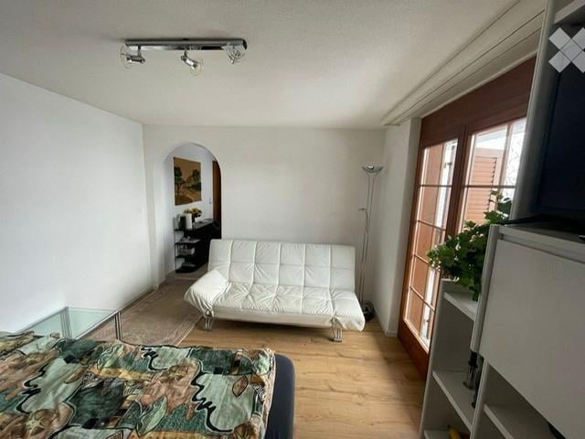 A modern, open-concept living and sleeping area with a white sofa, a bed with a colorful bedspread, a glass coffee table, a floor lamp, and a shelf with decorative items.