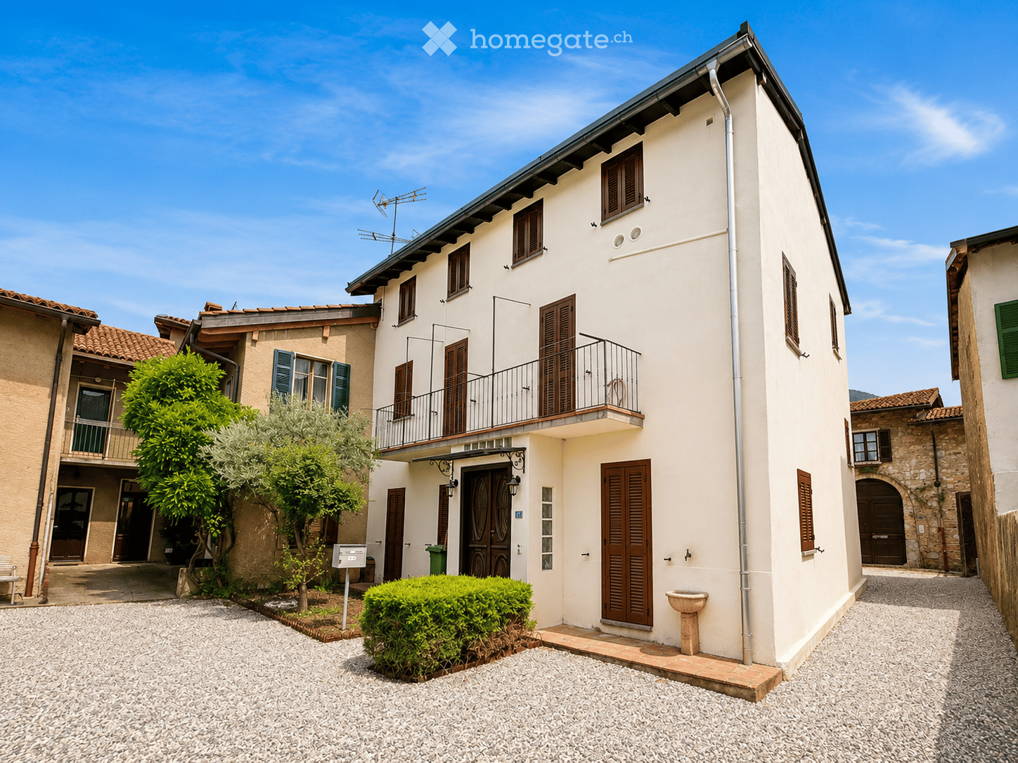Two-story house with beige walls, wooden shutters, and a balcony on the upper floor. The house features a gravel driveway, small garden area with shrubs and trees, and a stone fountain near the entrance.