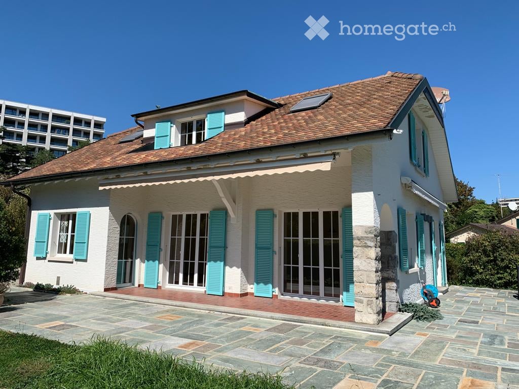 Two-story house with white exterior, teal shutters, and a tiled roof. Features a covered entrance and a paved patio area.