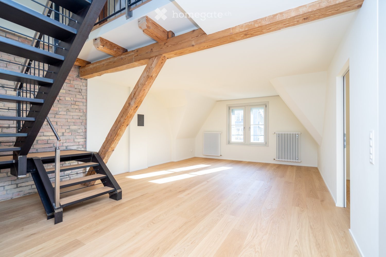Empty room with wooden flooring, exposed wooden beams, and a staircase leading upwards. The walls are painted in a light color, and there are radiators on either side of a window.