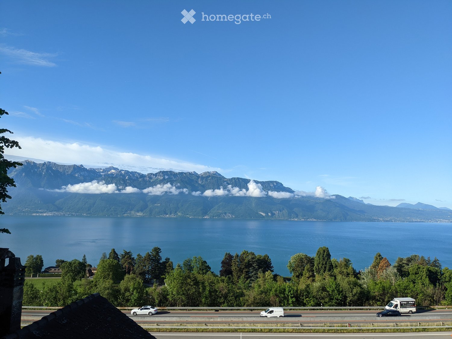 Scenic view of a lake surrounded by mountains, with a road running alongside and a few vehicles visible.