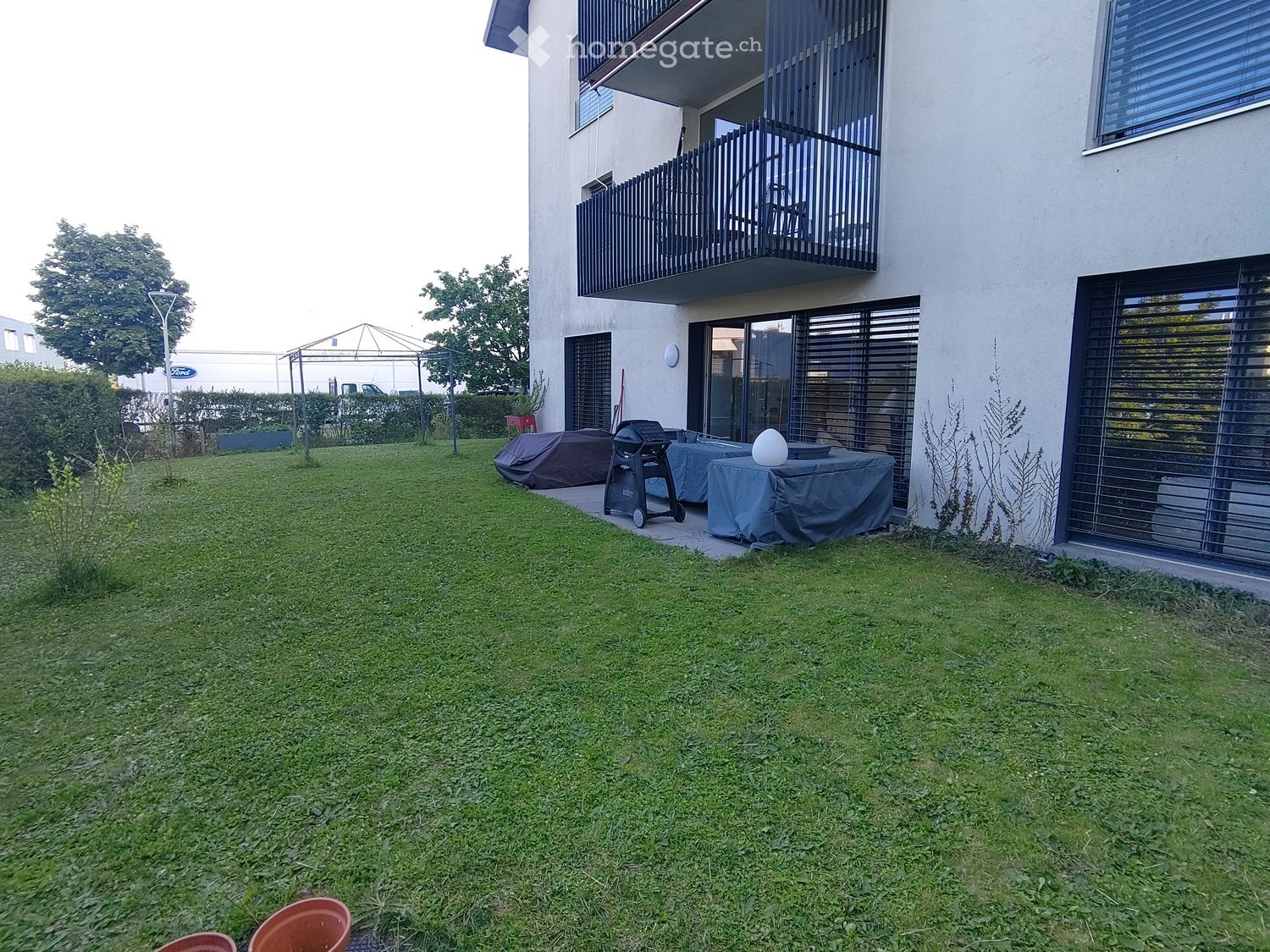 Well-maintained lawn with a patio area featuring a covered table, grill, and chairs, adjacent to a modern building with a balcony.