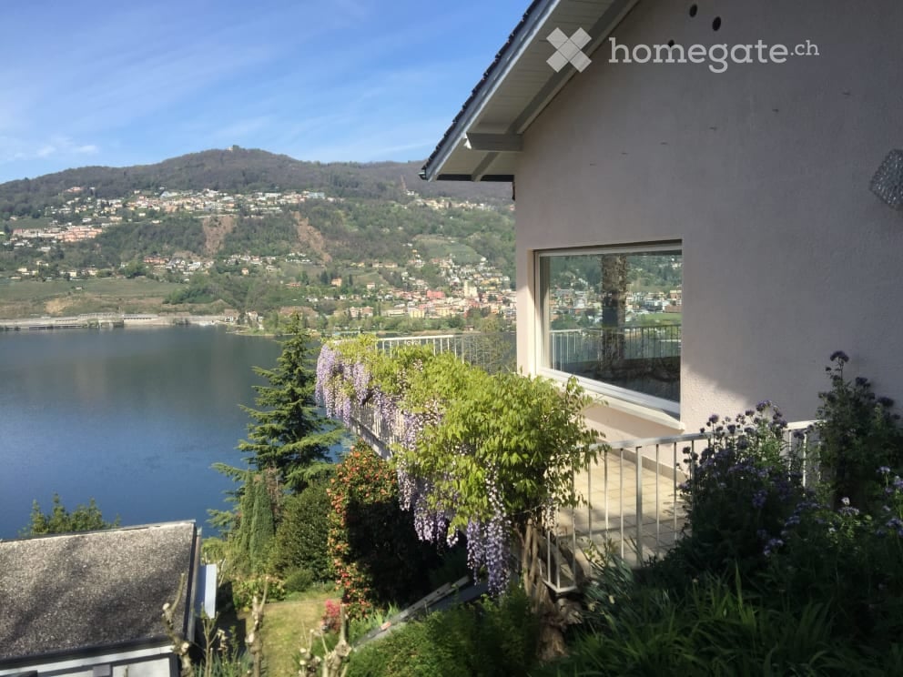 Balcony with a scenic view of a lake and surrounding hills, decorated with flowering plants and a small table.
