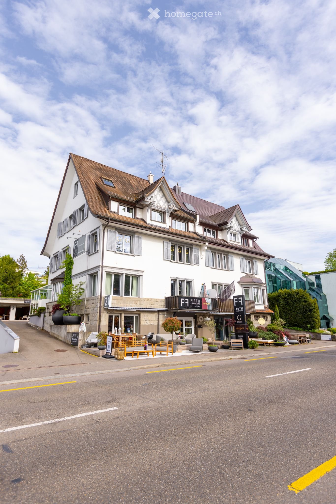 Three-story white building with brown roof, multiple windows, ground floor shop with outdoor seating, several plants and furniture, yellow line marking lane