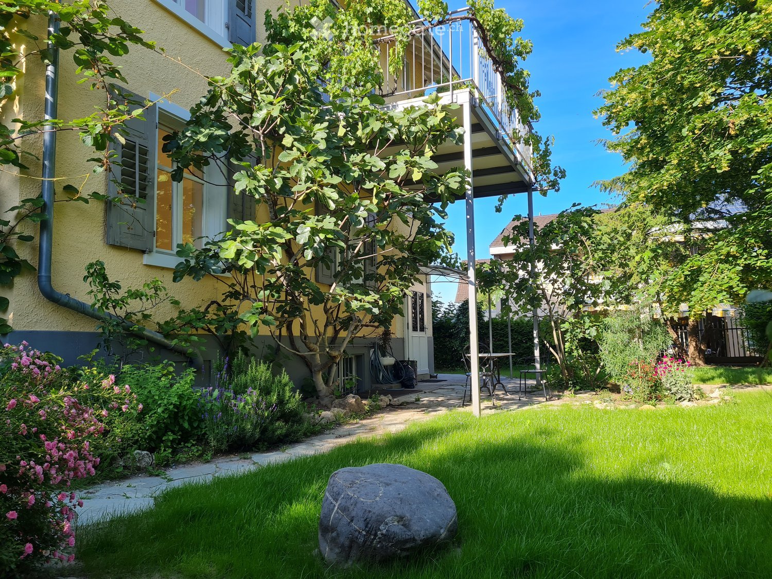 A small garden with a green lawn, trees, and a balcony attached to the house