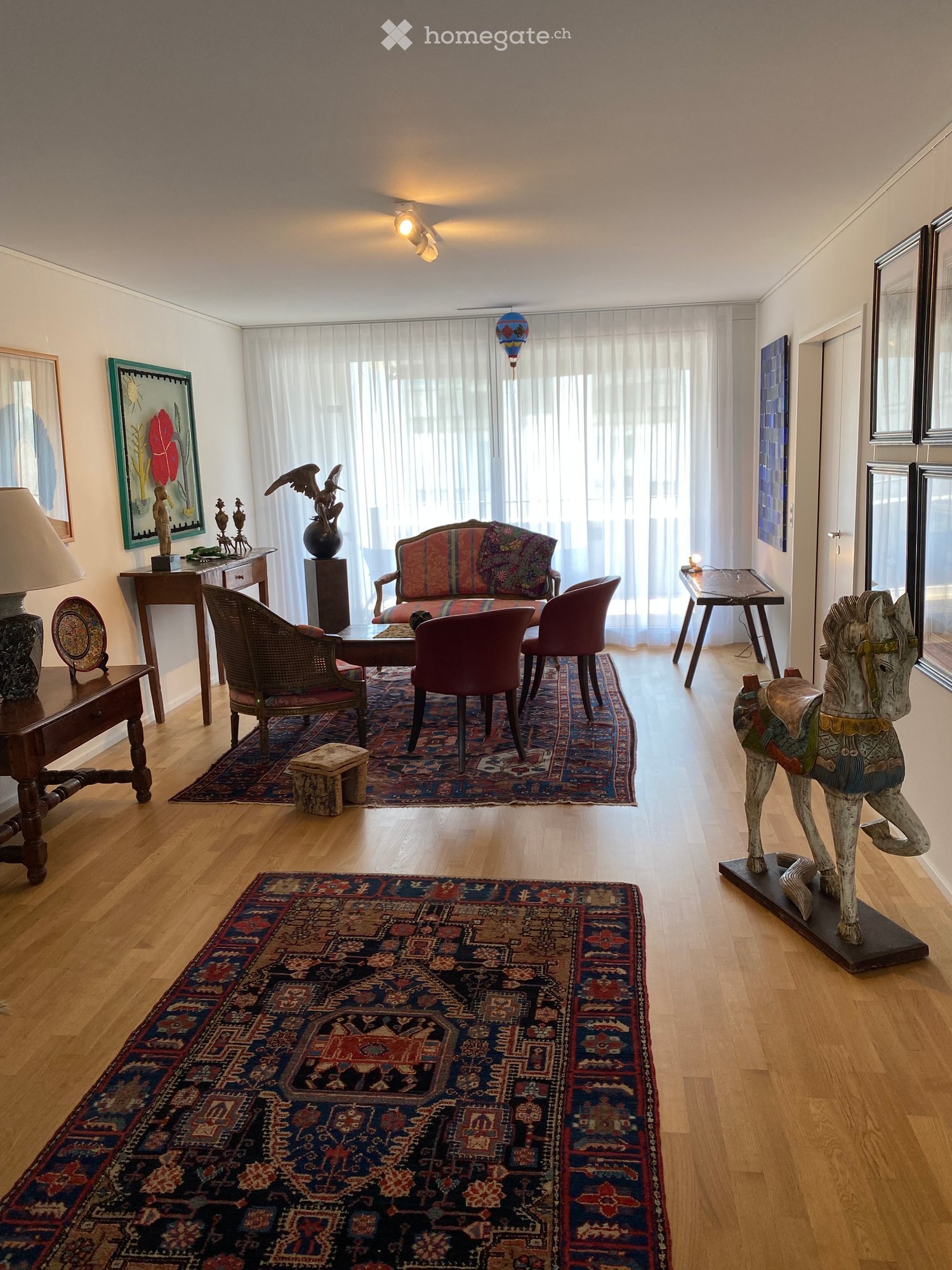 Living Room. Elegant living room with hardwood floors, large windows with sheer curtains, colorful rugs, a patterned sofa, armchairs, a coffee table, side tables, lamps, and decorative sculptures.
