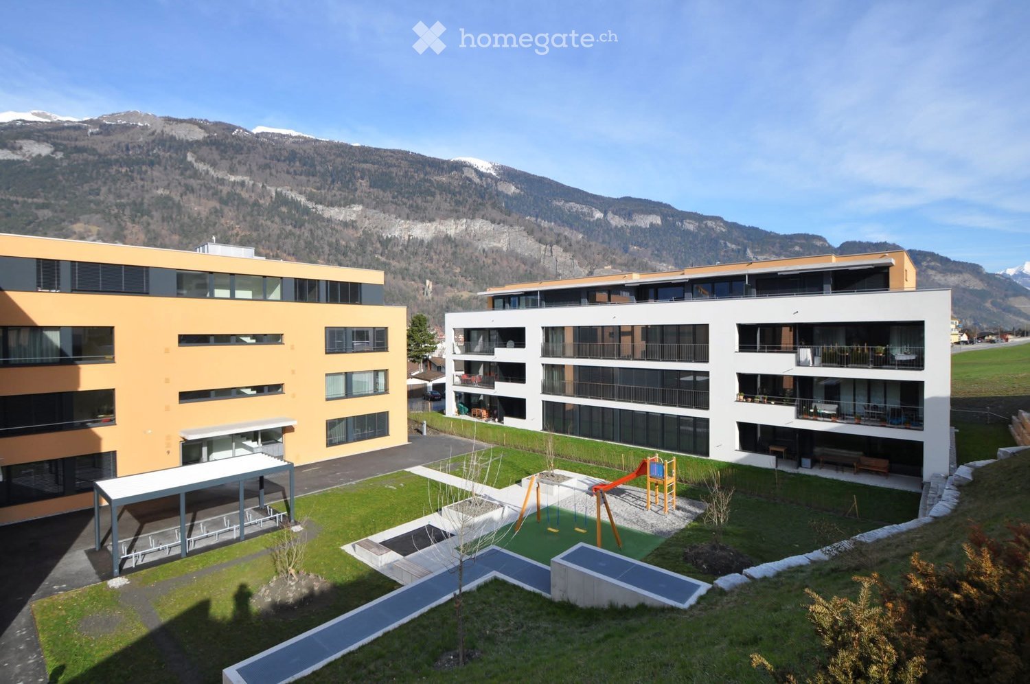 Modern multi-story residential building with balconies, surrounded by a landscaped garden area featuring a playground, small water features, and seating areas.