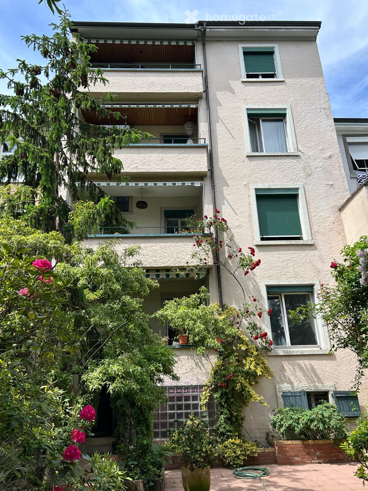 Four-story residential building with balconies, green shutters, and surrounding garden with potted plants and flowers.