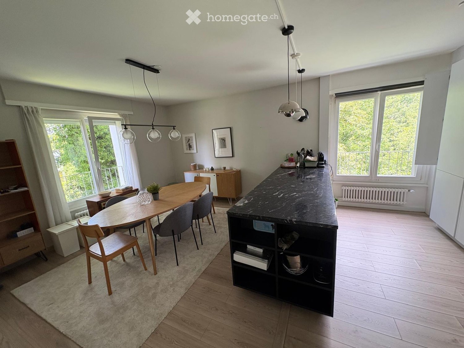 Open-plan dining and kitchen area with wooden dining table, chairs, kitchen island, pendant lights, and large windows overlooking a balcony.