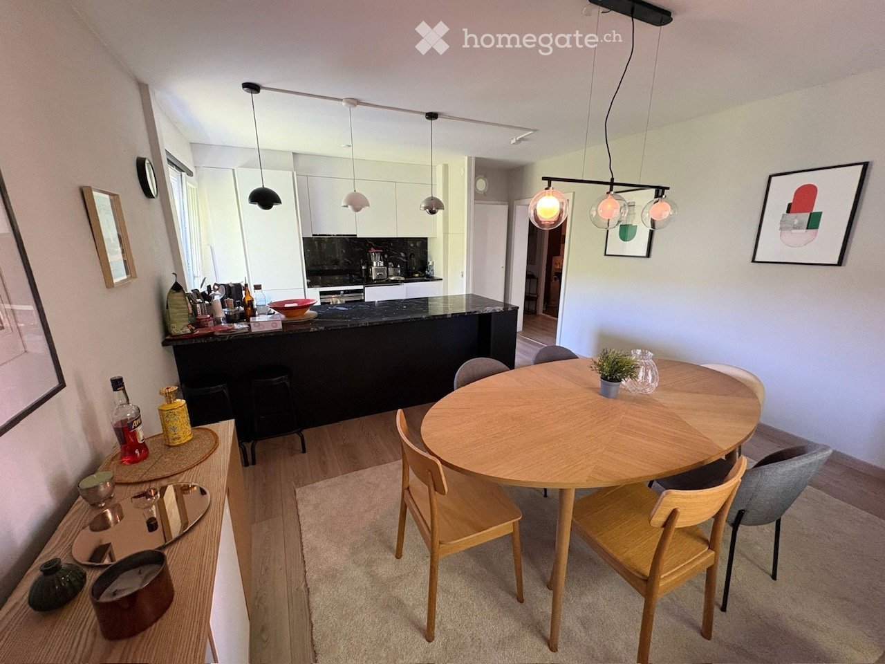 A modern, open-concept living and dining area with a round wooden dining table, chairs, a kitchen island, and pendant lighting. The space features a sleek black countertop, various kitchen items, and decorative elements like framed artwork and a small pla