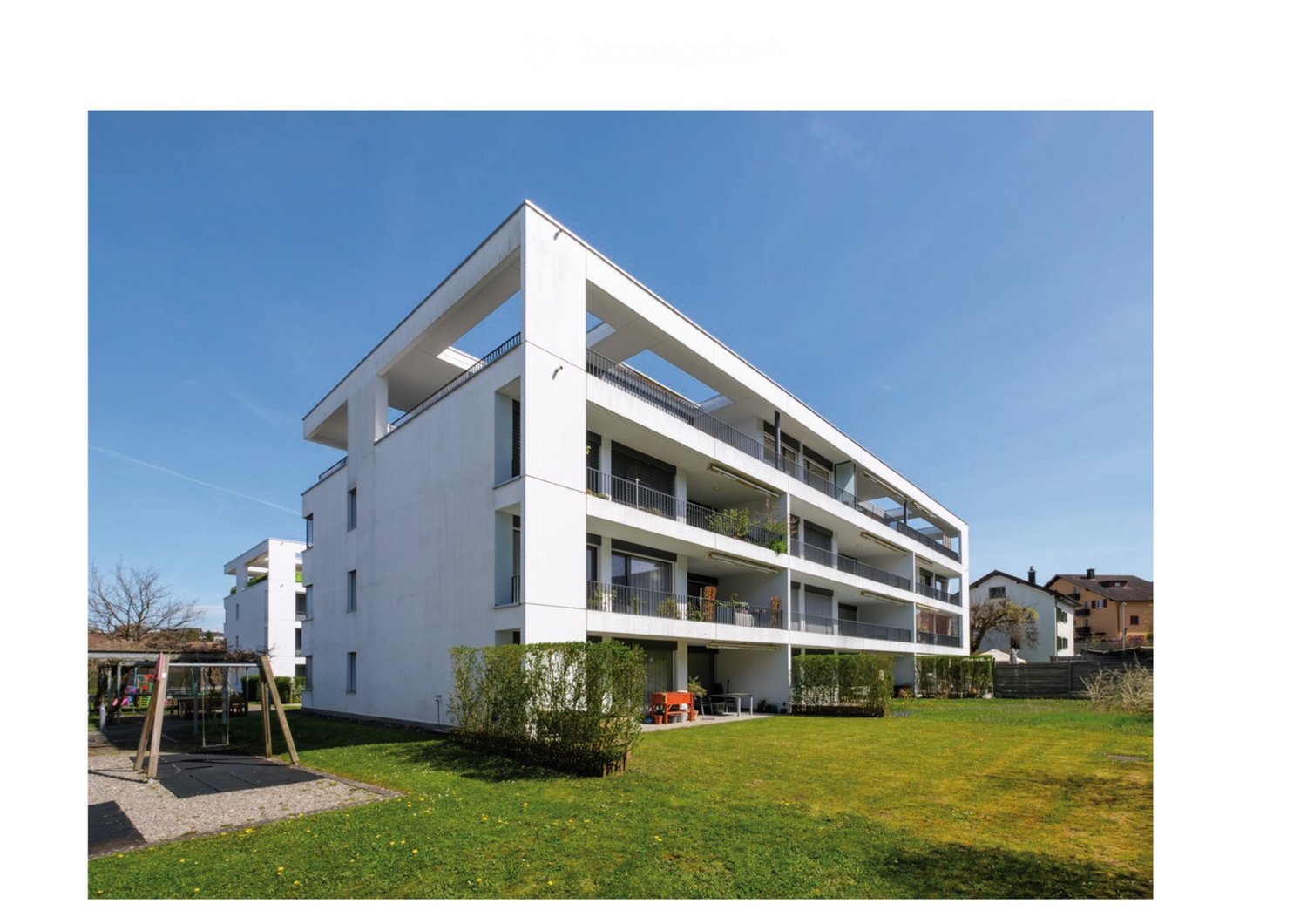 Modern multi-story residential building with white facade, balconies, and surrounding green lawn.