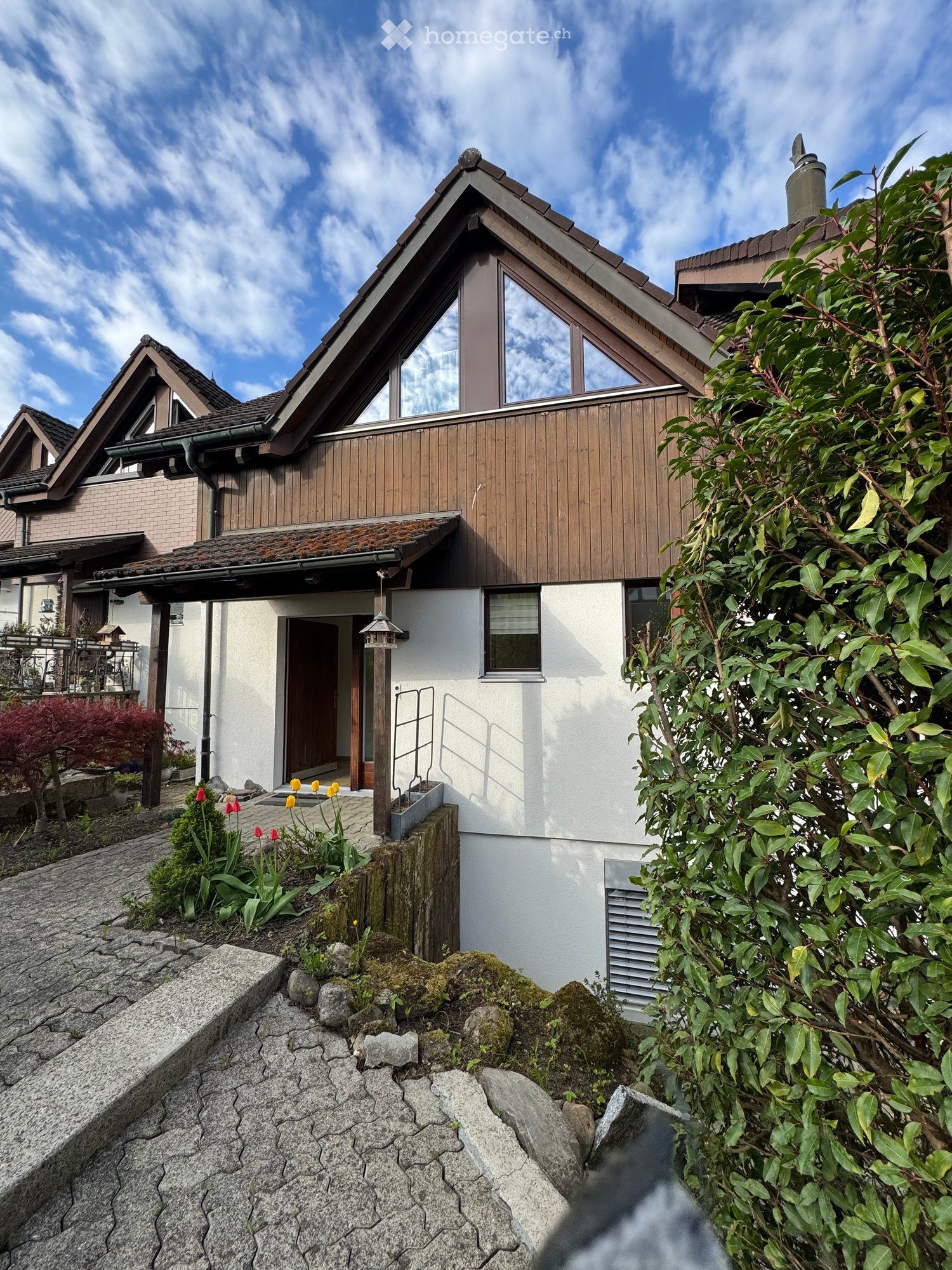 Two-story house with a wooden facade, white walls, and a sloped roof. Features a small garden with flowers and rocks in the foreground.