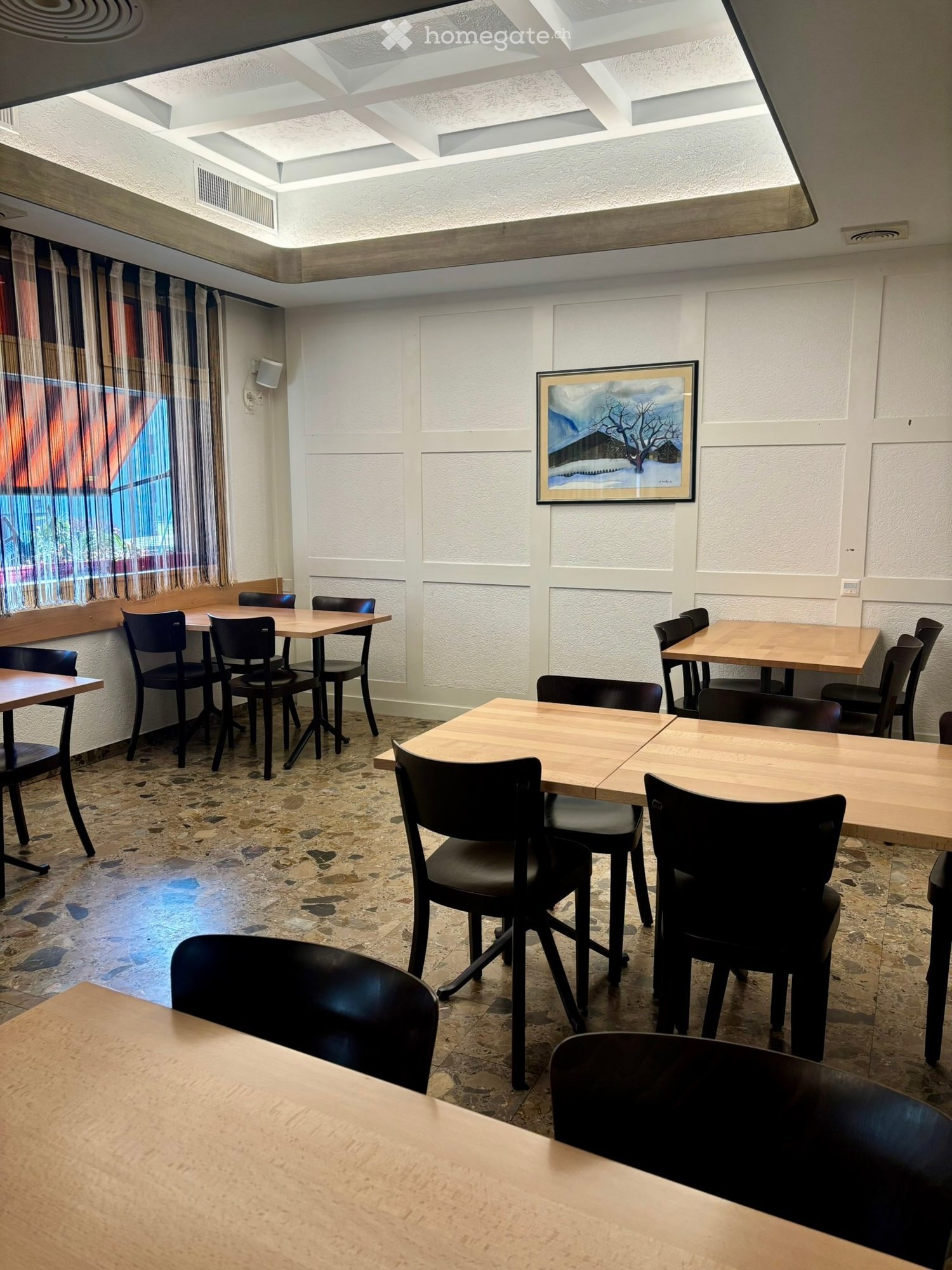 A spacious dining area with multiple wooden tables and black chairs arranged throughout, featuring a large painting on the wall and a large window with curtains.