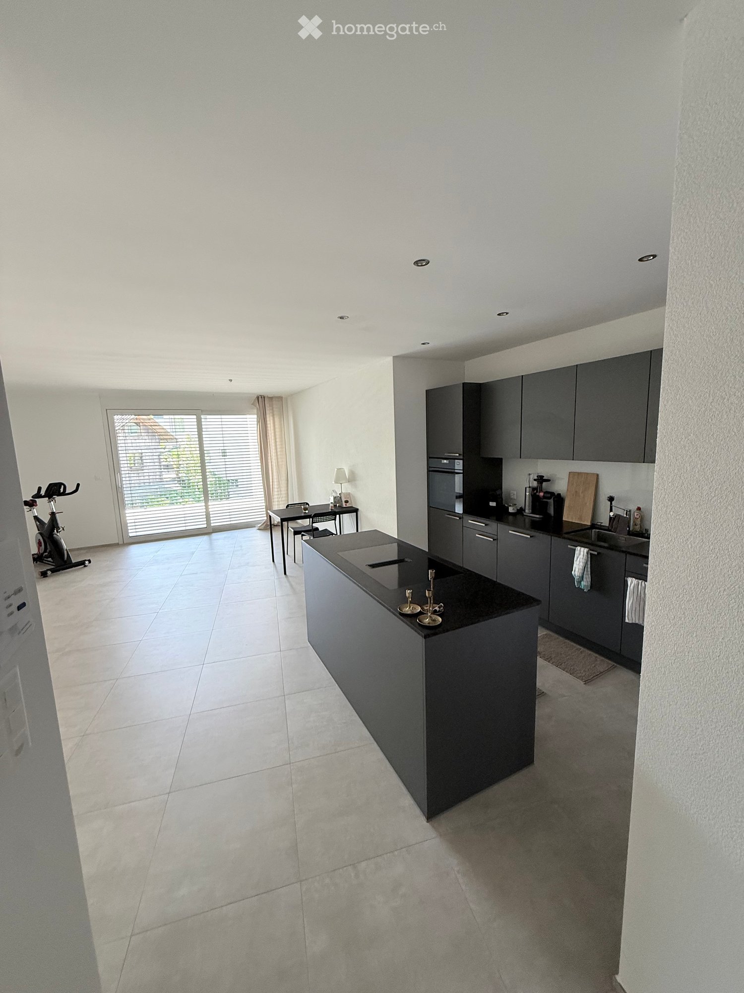 Modern kitchen with grey cabinets, a central island, a sink, and a small dining area with a table and chairs. An exercise bike is positioned near the window.