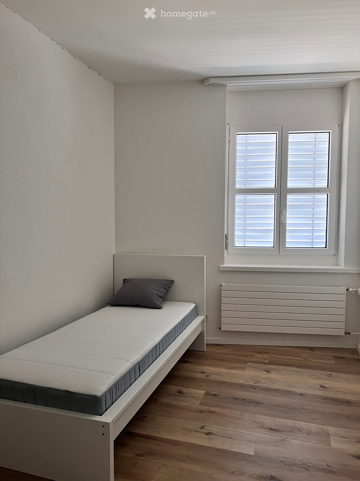 Single bed, white headboard, grey pillow, white walls, wooden floor, window with white shutters, white radiator