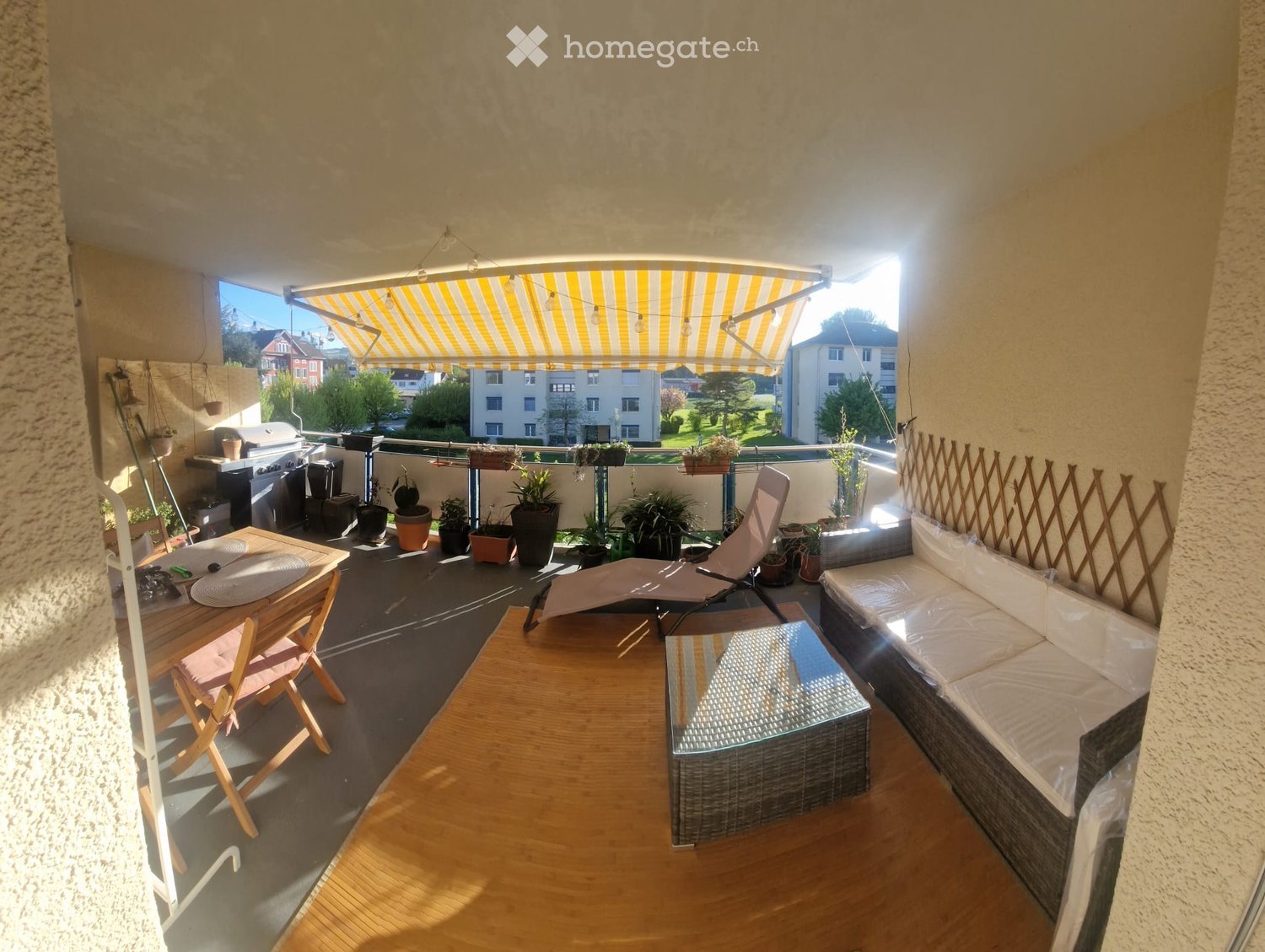 Balcony with a wooden floor, a covered seating area, a table with chairs, a sofa, a lounge chair, and several potted plants.