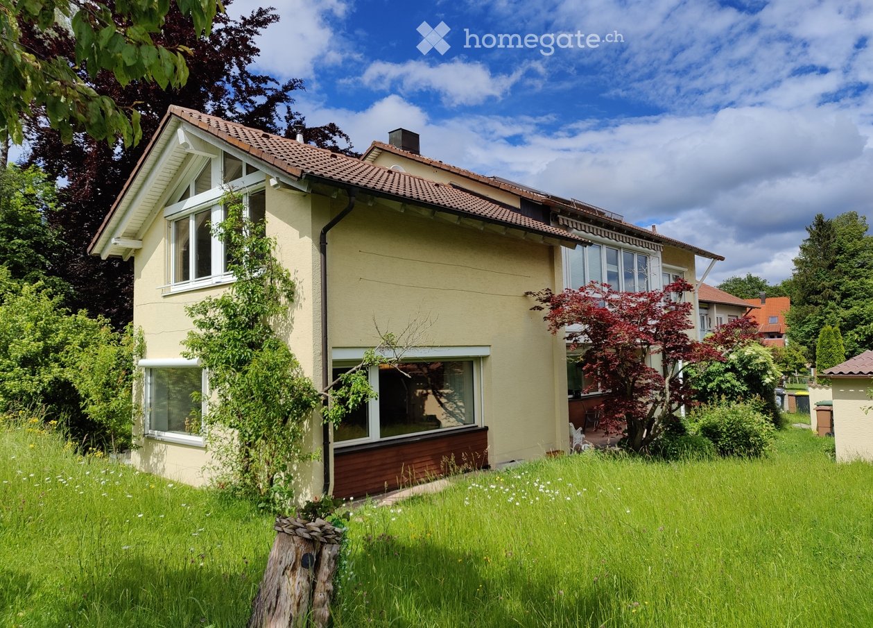 Two-story house with beige walls, red-tiled roof, multiple windows, and a well-maintained garden with trees and shrubs.