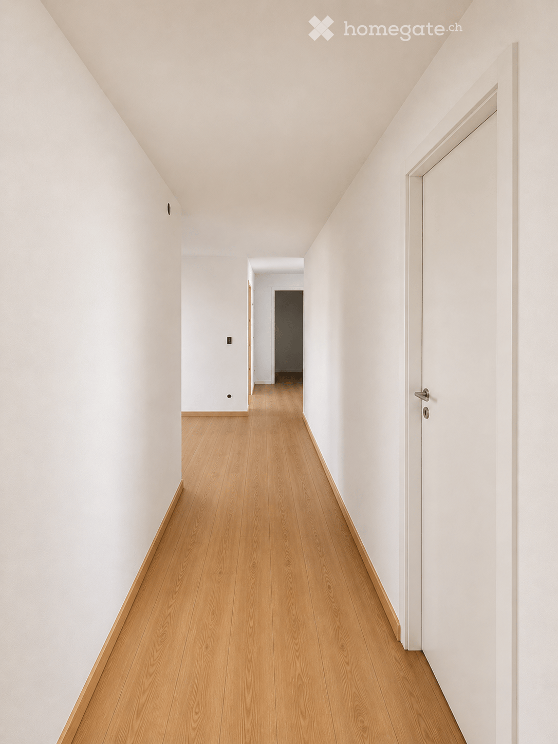Minimalist hallway with light wooden flooring and white walls, featuring a closed door at the end and a visible light switch on the left wall.