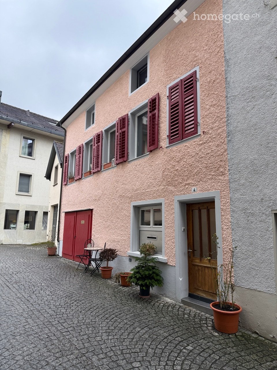 Pink and grey building, red shutters, wooden door, small table with chairs, multiple potted plants