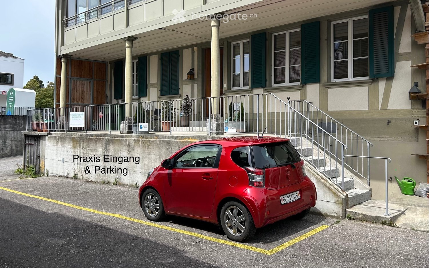 A two-story building with a balcony, green shutters, and a staircase leading up. A red car is parked in front, and there is a sign indicating 'Praxis Eingang & Parking'.