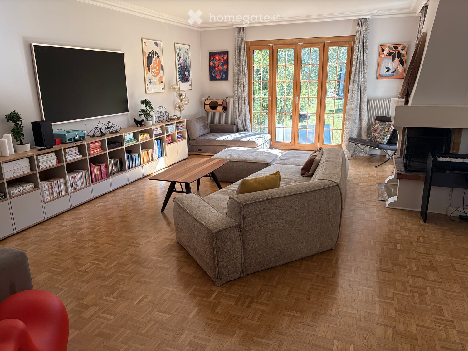 Modern living room with herringbone wooden flooring, large sectional sofa, coffee table, bookshelf, fireplace, and large TV screen.