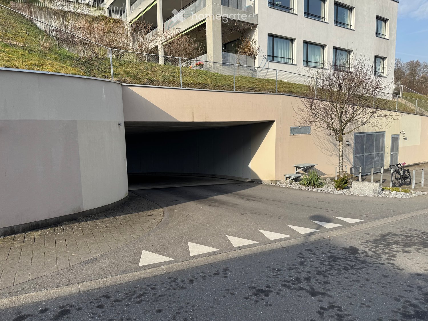 Modern multi-story building with a raised ground floor, garage entrance, and a small landscaped area with a picnic table and bicycle racks.