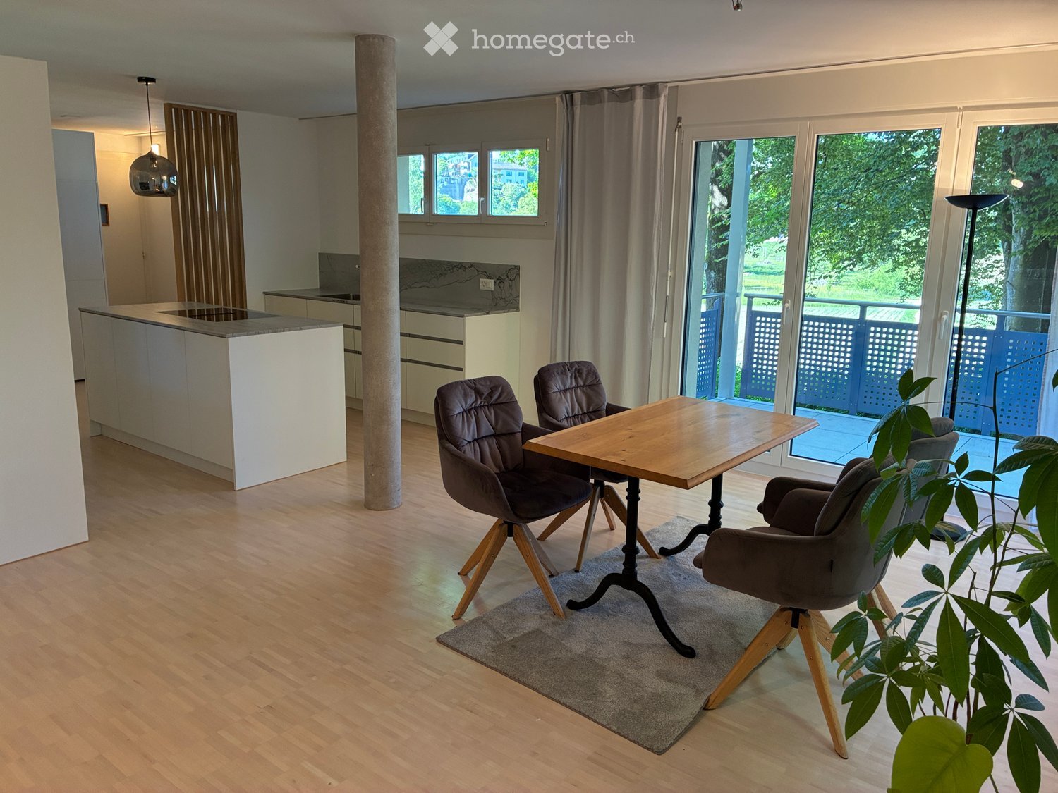 Modern open-plan living and dining area with wooden flooring, a central dining table with four chairs, a kitchen island, pendant lighting, and large windows leading to a balcony.