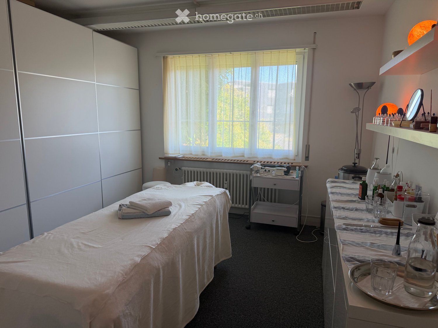 A well-lit room with a massage bed, shelves with various items, a cabinet, and a small table with cups and bottles.