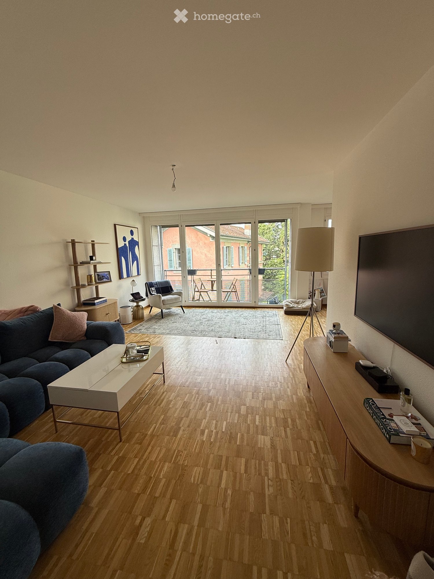 Modern living room with wooden flooring, large sliding glass doors leading to a balcony, a blue sectional sofa, a white coffee table, an armchair, a floor lamp, and a TV stand.