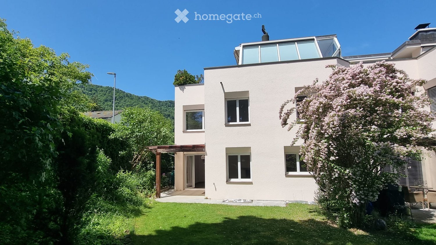 Modern two-story house with white exterior, large windows, and a flat roof terrace. The house is surrounded by a well-maintained garden with a pergola-covered entrance and blooming trees.