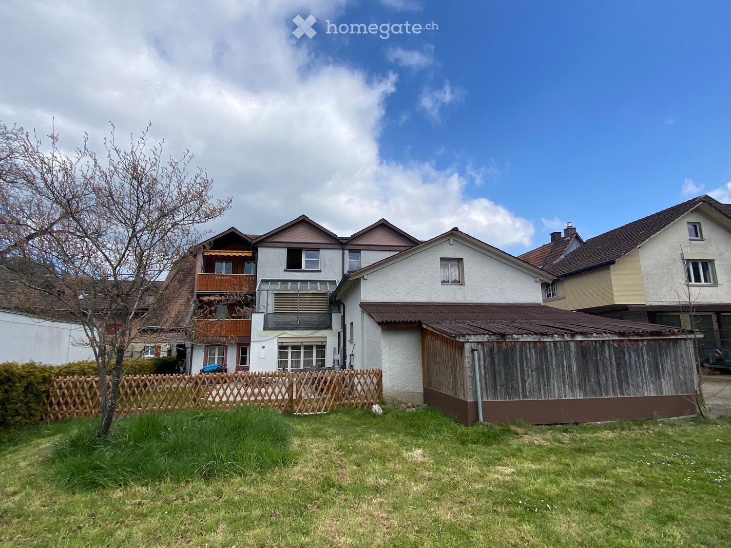 Two-story house with white walls, brown roof, and balcony. The house is surrounded by a wooden fence and a small garden area.