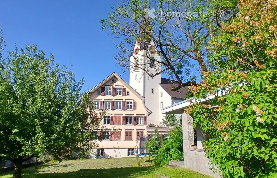 A two-story building with a tower, surrounded by trees and greenery, with multiple windows and a sloped roof.