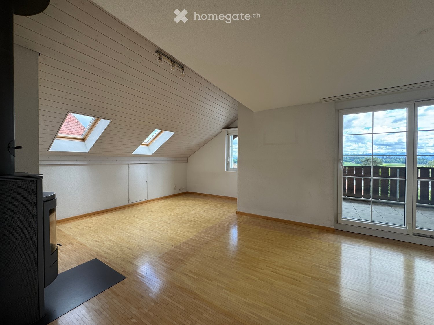 Bright, empty attic room with wooden floors, slanted wooden ceiling, skylights, and large sliding glass doors leading to a balcony.