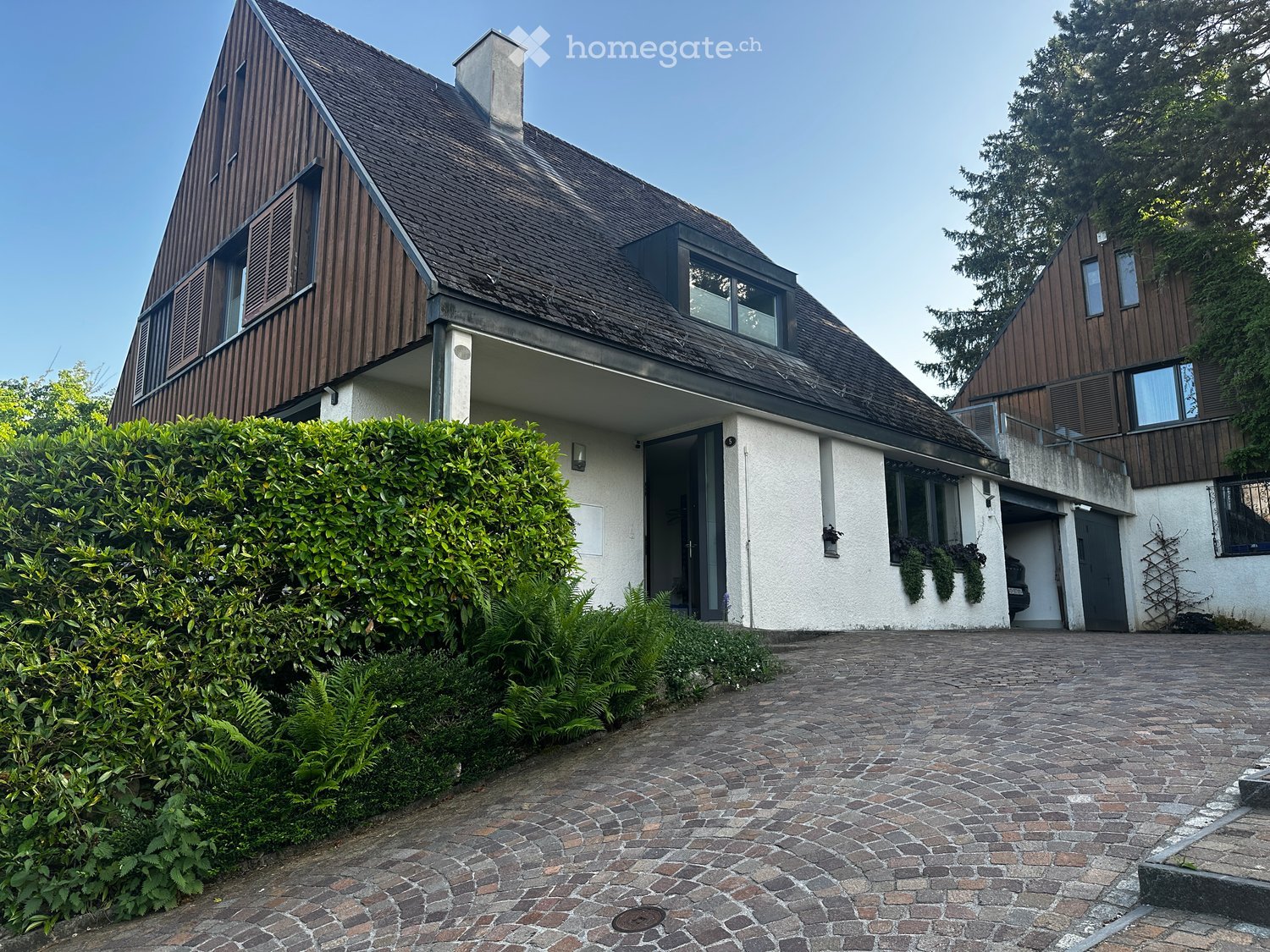 Two-story house with a modern design, featuring a wooden facade, large windows, and a well-maintained garden with a paved driveway.