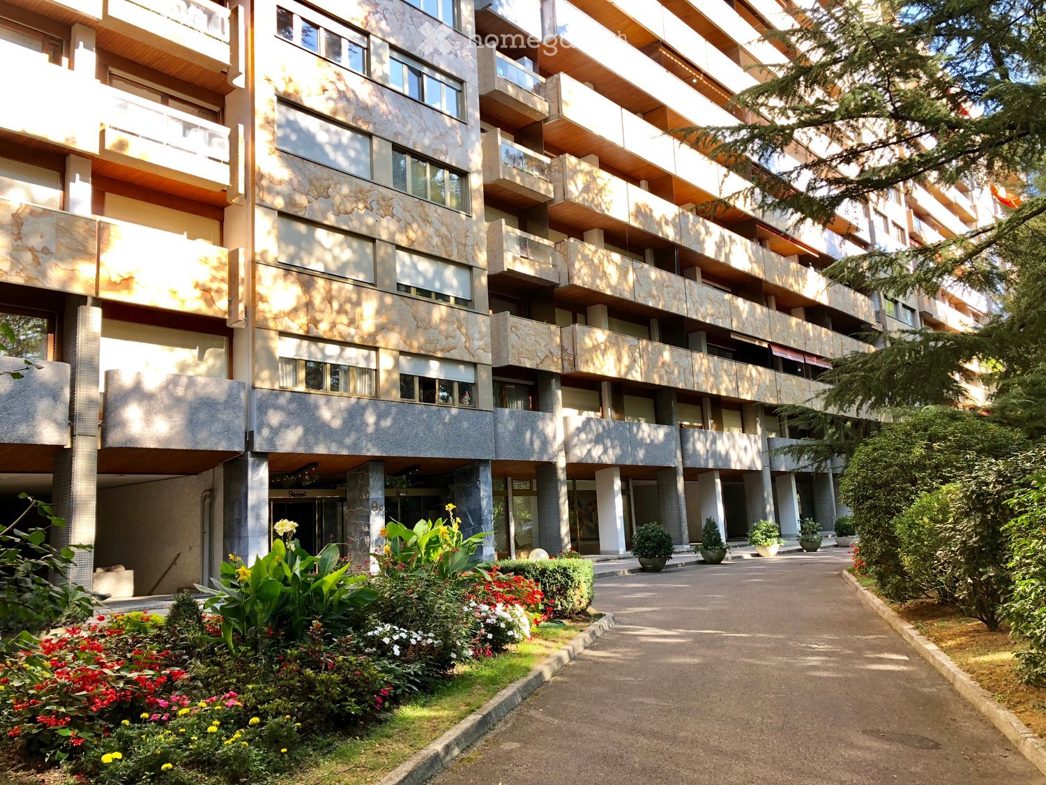Multi-story apartment building with balconies, stone facade, and landscaped garden area with flowers and shrubs.