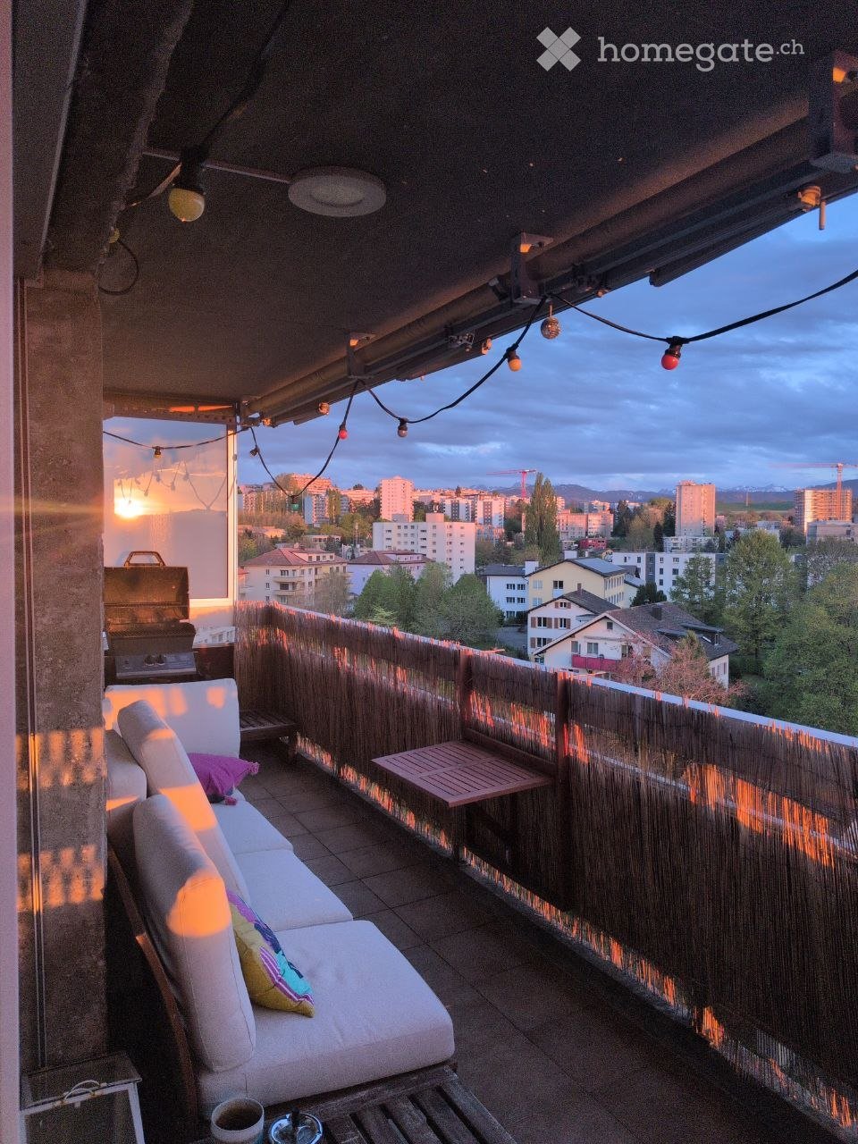 Balcony with a white sofa, small table, grill, and string lights, overlooking a cityscape at sunset.